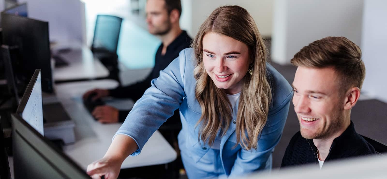Twee collega’s werken samen achter een computer terwijl een derde collega op de achtergrond aan het werk is.