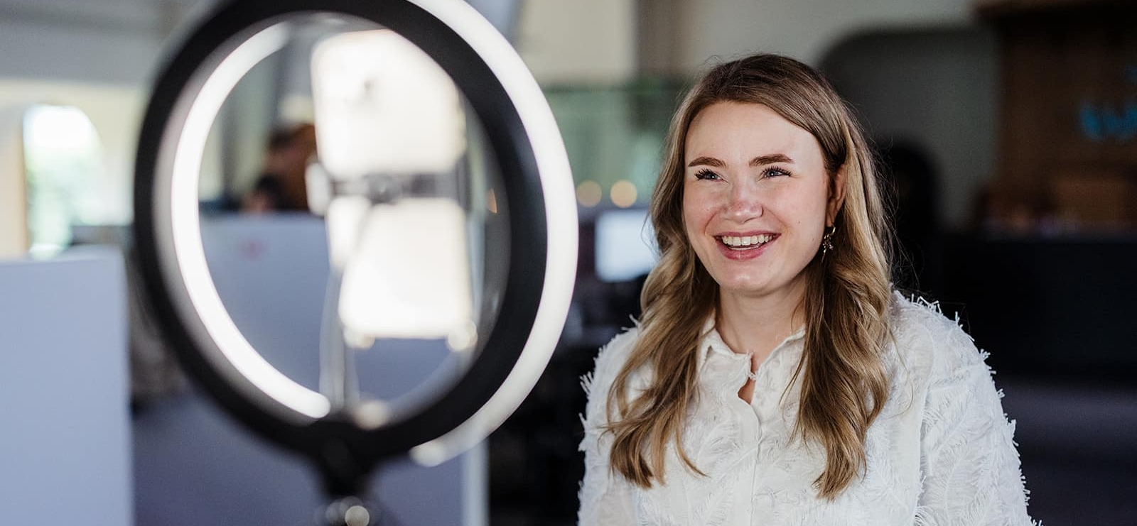 Vrouw die glimlachend voor een ringlamp zit, alsof ze een video opneemt in een kantooromgeving.