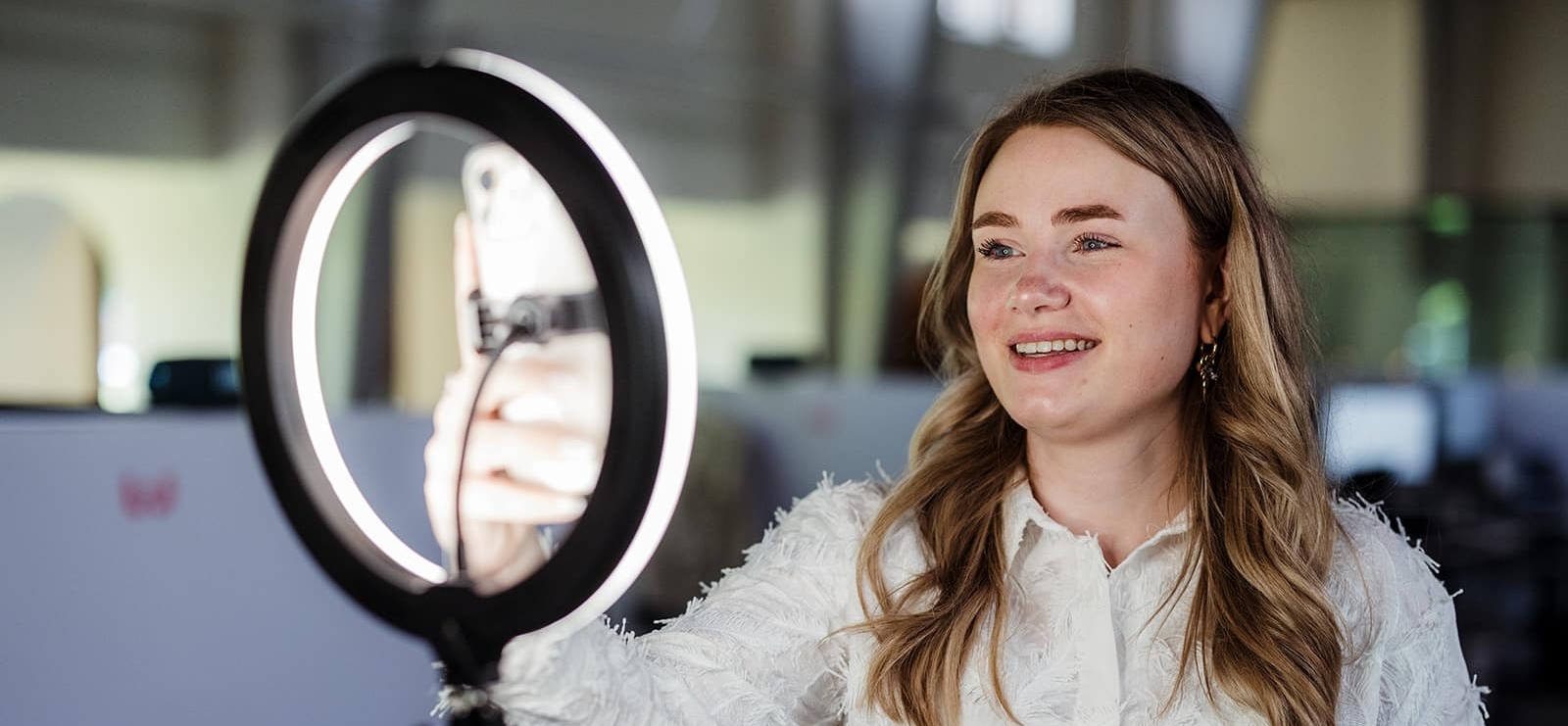 Vrouw maakt een selfie of video met smartphone voor een ringlamp in een kantooromgeving.