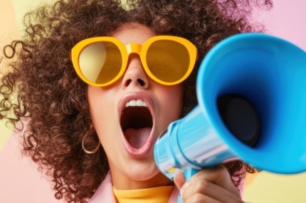 Vrouw met krullend haar en gele zonnebril roept door een blauwe megafoon tegen een kleurrijke achtergrond.