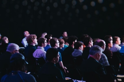 Publiek dat aandachtig luistert tijdens een presentatie of conferentie in een donkere zaal.