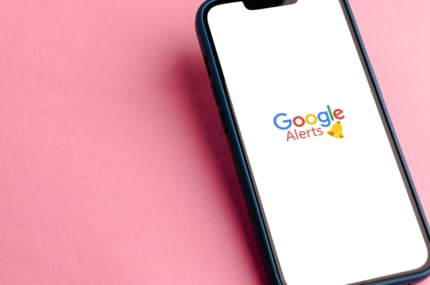 Smartphone met het Google Alerts-logo op het scherm tegen een roze achtergrond.