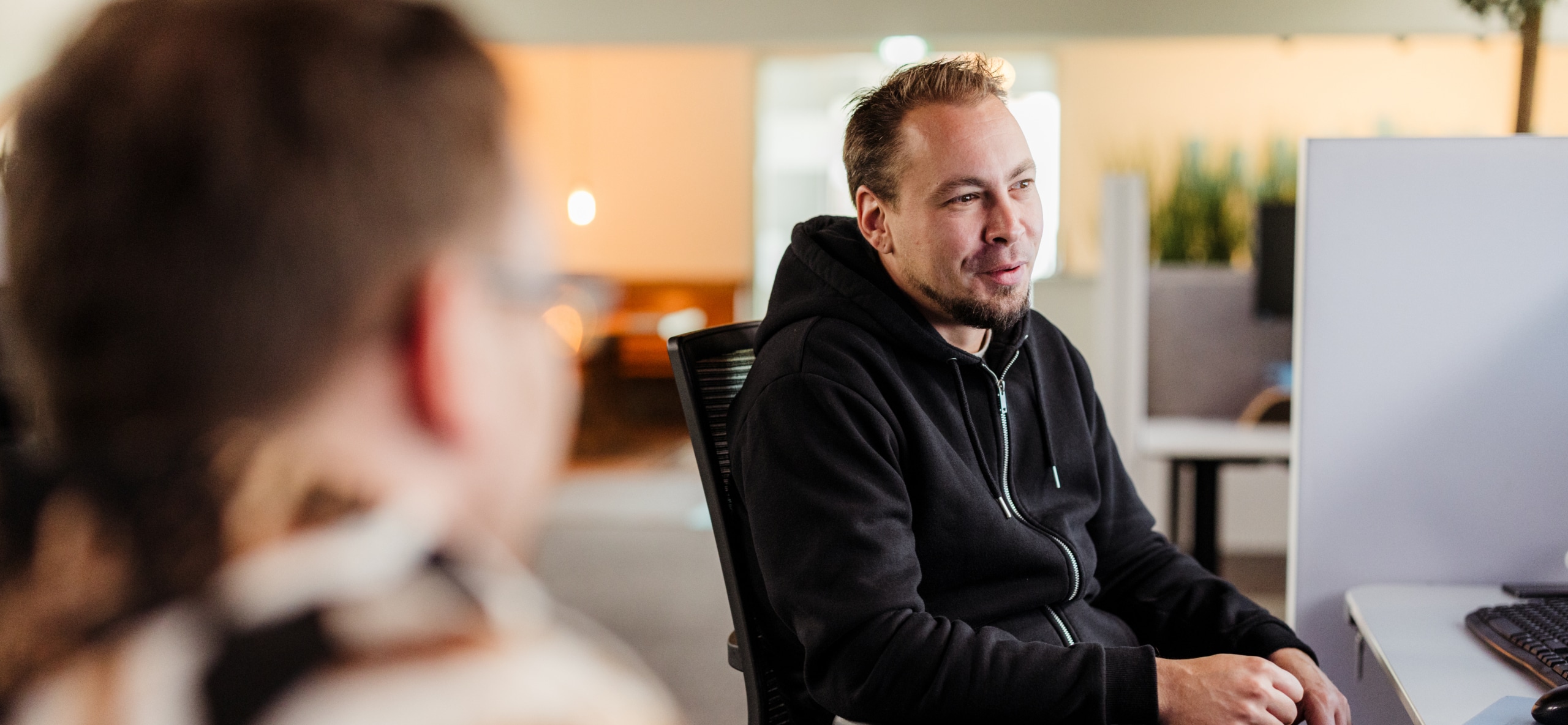 Man in een zwart vest in gesprek op kantoor, met een collega onscherp op de voorgrond.