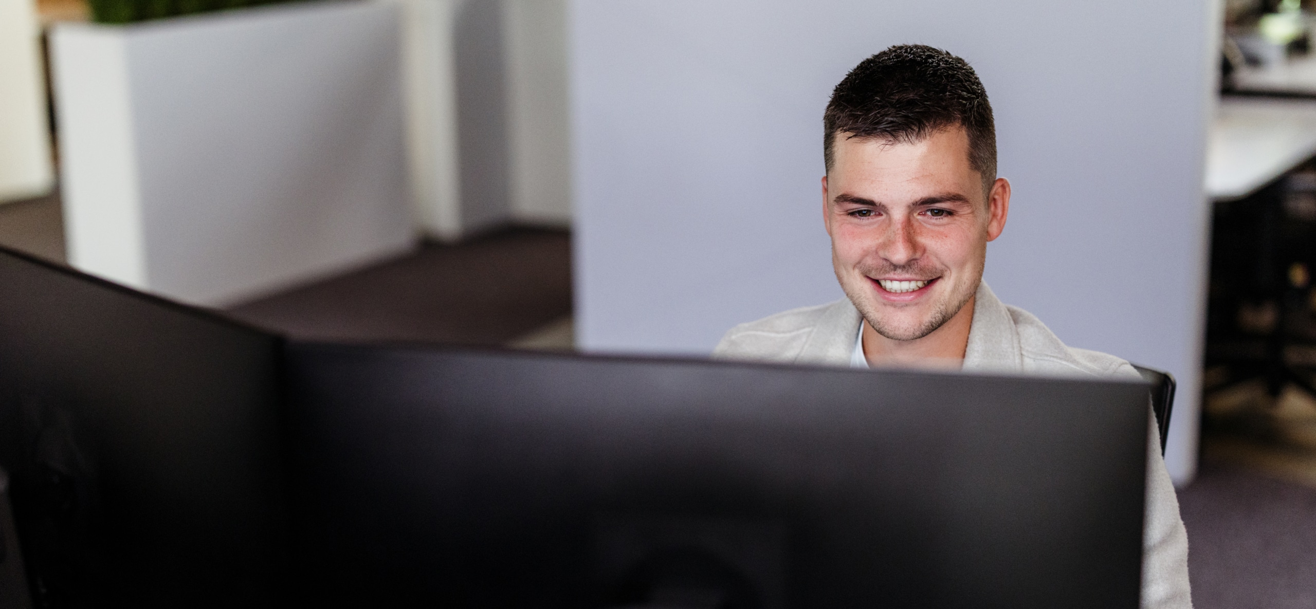 Een medewerker glimlacht terwijl hij achter een groot computerscherm werkt in een kantooromgeving.