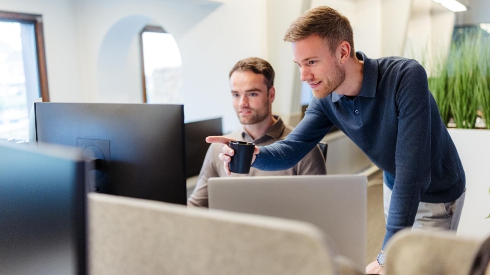Twee medewerkers werken samen aan een bureau in een kantooromgeving. Een man wijst naar het scherm van zijn collega, terwijl deze geïnteresseerd naar de computer kijkt. Beide zijn in casual kleding en gefocust op het scherm.