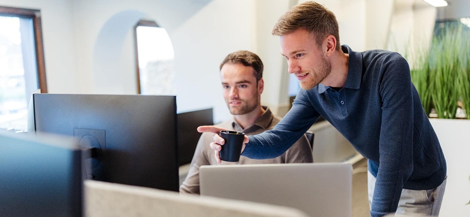 Twee collega’s werken samen achter een bureau en kijken naar een computerscherm, waarbij één man naar het scherm wijst.