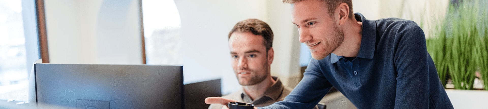 Twee collega’s werken samen achter een bureau en kijken naar een computerscherm, waarbij één man naar het scherm wijst.
