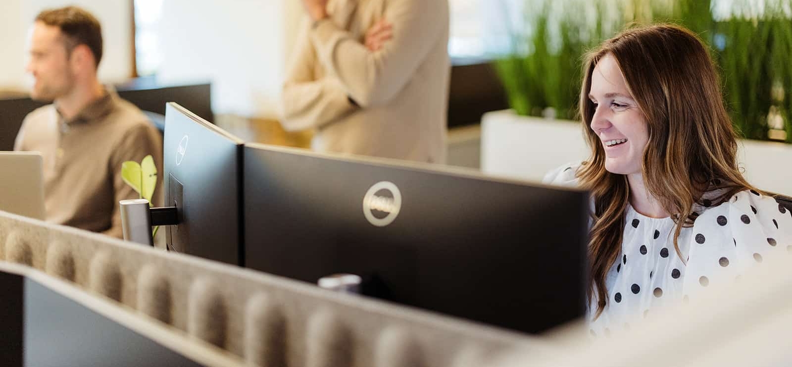 Een medewerker glimlacht terwijl ze achter twee computerschermen werkt in een moderne kantooromgeving.
