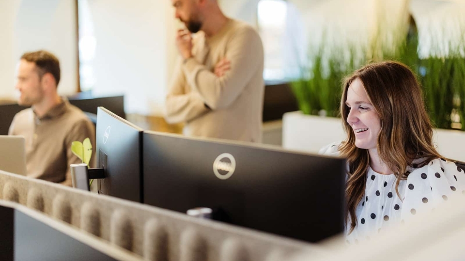Een vrouw lacht terwijl ze achter haar bureau zit in een kantoorruimte, terwijl een man in de achtergrond ook aan zijn werkstation zit. Het kantoor is goed verlicht en er staan planten op de bureaus.