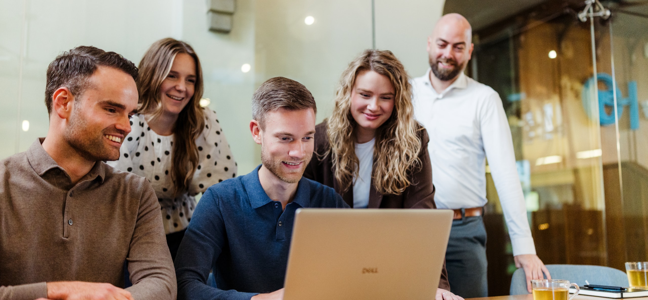 Een team van vijf mensen in een vergaderruimte kijkt naar een laptop. Ze zijn gefocust op de gegevens op het scherm, met een ontspannen en gemotiveerde sfeer.