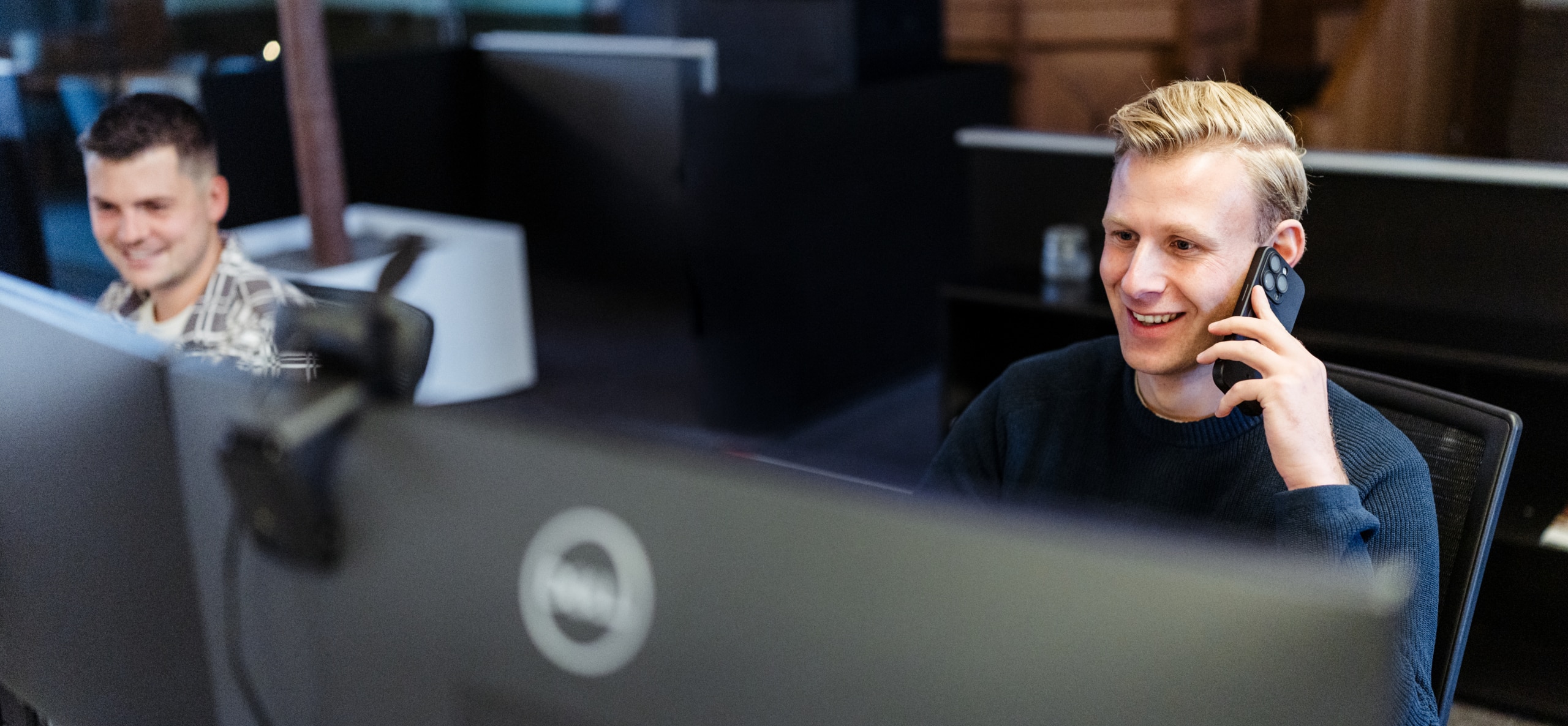 Een man zit achter zijn bureau in een kantoor en belt met zijn smartphone, terwijl een collega op de achtergrond achter een computer werkt.