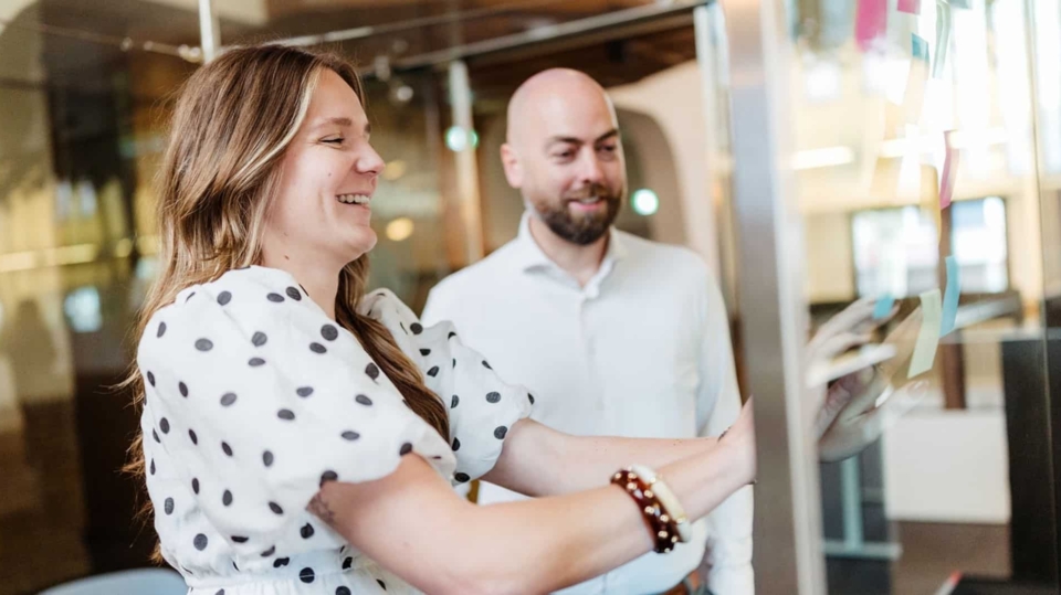 Een man en een vrouw glimlachen naar elkaar in een gesprek, terwijl de man een lichte glimlach toont en zijn hand opheft in een informele houding.