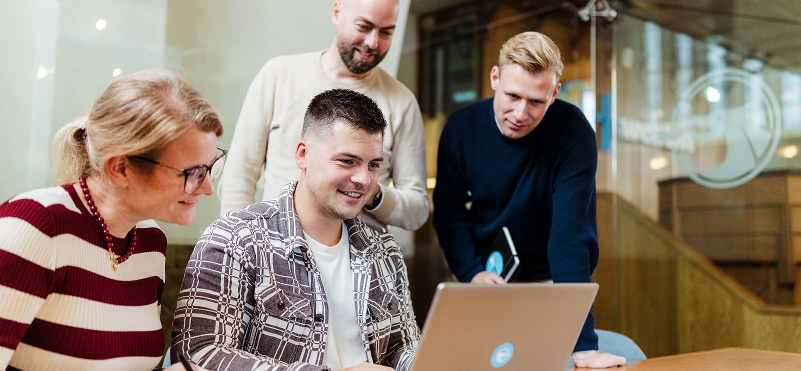 Vier medewerkers staan rond een laptop en lachen terwijl ze samenwerken. Een van hen kijkt naar het scherm, terwijl de anderen aandachtig meekijken.