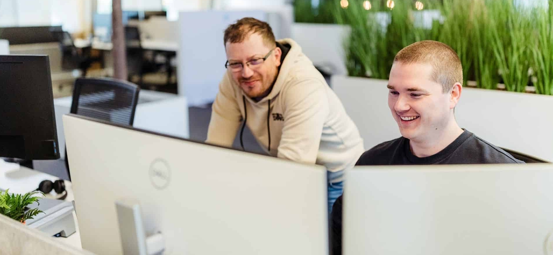 Twee collega’s werken samen achter computers in een kantoor en glimlachen terwijl ze naar het scherm kijken.