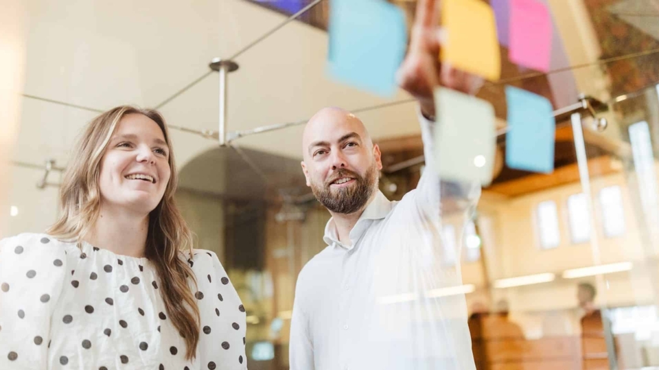 Een vrouw en een man staan in een vergaderruimte en kijken naar een reeks kleurrijke post-its die op een glazen wand zijn geplakt. De vrouw glimlacht terwijl de man wijst naar een post-it.