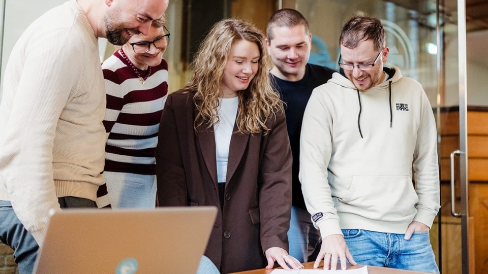 Een groep van zes mensen staat rond een tafel, met een laptop erop. Ze kijken gezamenlijk naar documenten op de tafel, terwijl ze met elkaar discussiëren. De sfeer is informeel en samenwerkingsgericht.