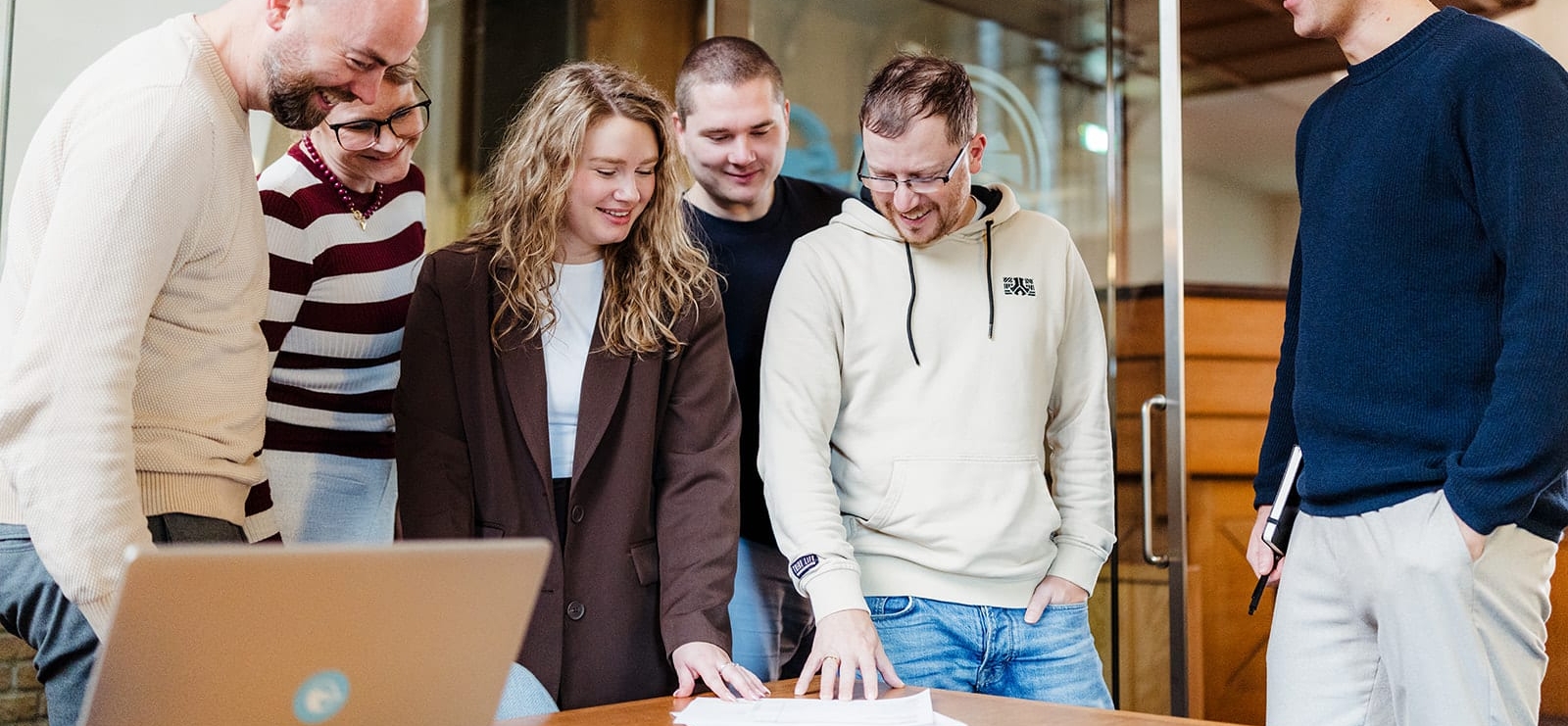 Een groep van zes mensen staat rond een tafel, met een laptop erop. Ze kijken gezamenlijk naar documenten op de tafel, terwijl ze met elkaar discussiëren. De sfeer is informeel en samenwerkingsgericht.