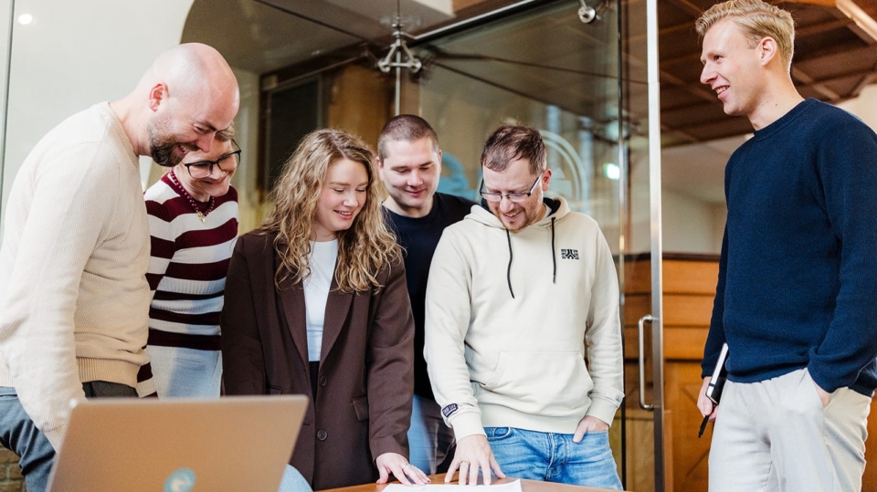 Een groep van zes mensen staat rond een tafel, met een laptop erop. Ze kijken gezamenlijk naar documenten op de tafel, terwijl ze met elkaar discussiëren. De sfeer is informeel en samenwerkingsgericht.