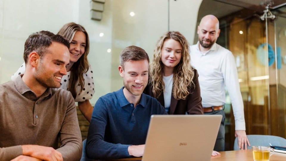 Vijf mensen staan rond een laptop die op een tafel staat. Ze kijken naar het scherm met interesse en lachen, terwijl ze samenwerken aan een project. De sfeer is informeel en samenwerkingsgericht.