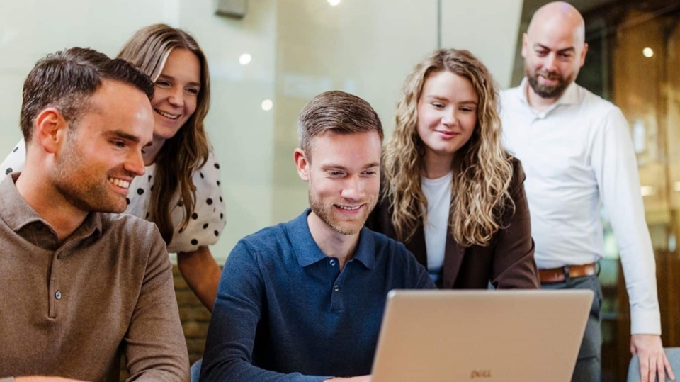 Vijf mensen staan rond een laptop die op een tafel staat. Ze kijken naar het scherm met interesse en lachen, terwijl ze samenwerken aan een project. De sfeer is informeel en samenwerkingsgericht.