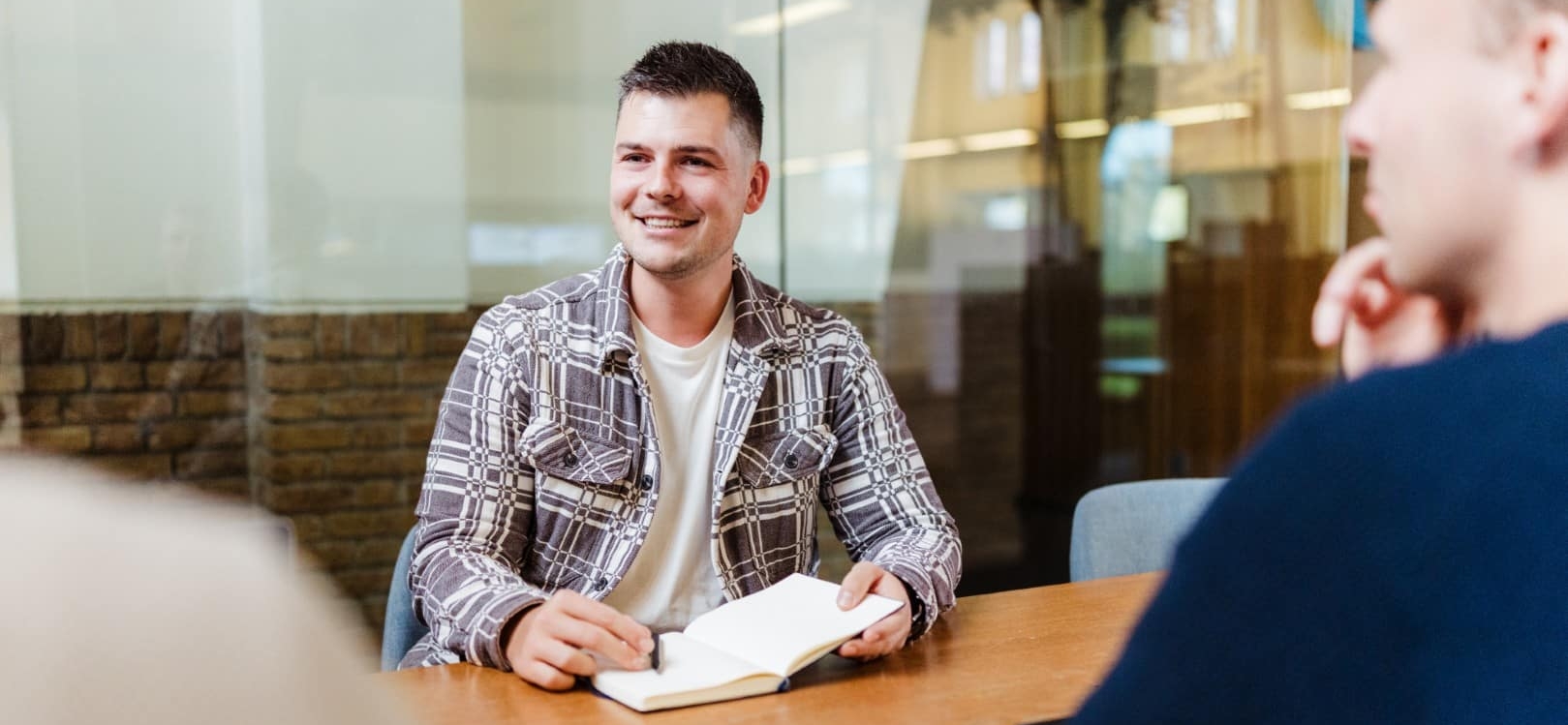 Een man zit aan een vergadertafel met een notitieboek en glimlacht terwijl hij in gesprek is met collega’s.