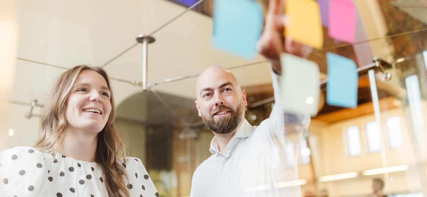 Een vrouw en een man staan samen in een vergaderruimte, beiden glimlachend. De man wijst naar een post-it terwijl de vrouw erbij staat en kijkt.