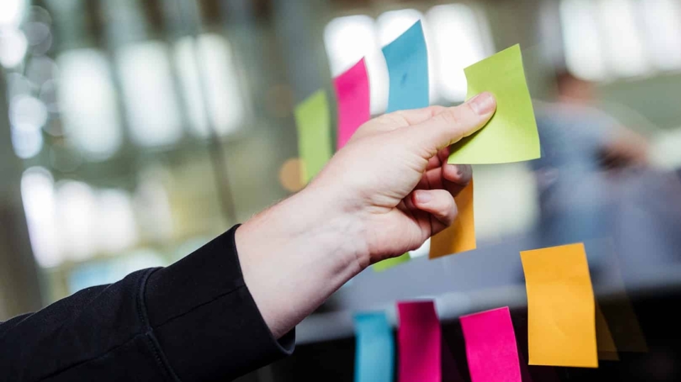 Een hand plakt gekleurde post-its op een glazen wand tijdens een brainstorm of overleg in een kantooromgeving.
