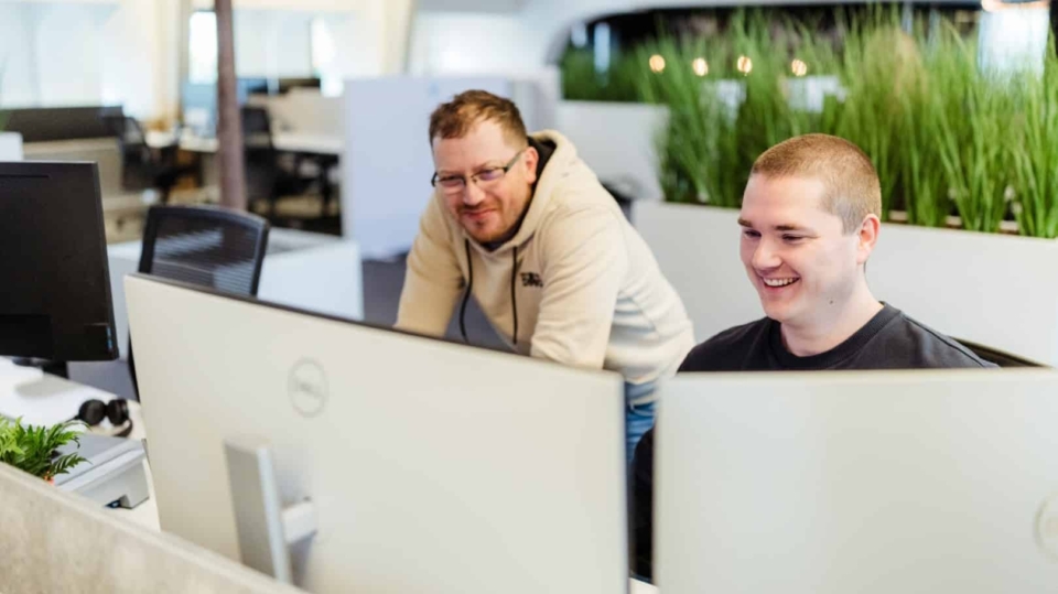 Twee collega’s werken samen achter computers in een kantoor, waarbij één man over de schouder meekijkt en beiden glimlachen naar het scherm.
