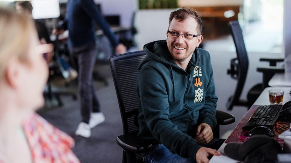 Een man glimlacht naar een collega terwijl ze aan hun werkstations zitten in een kantoor. Hij draagt een groen sweatshirt en lijkt een gesprek te hebben met de collega aan de andere kant van zijn bureau.
