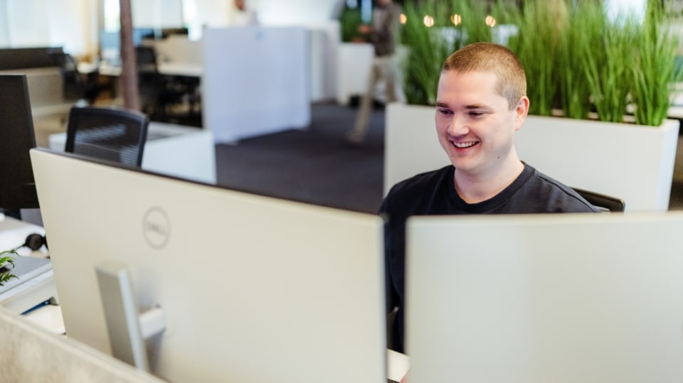 Een man glimlacht terwijl hij achter zijn bureau zit in een kantooromgeving, met planten op de achtergrond.