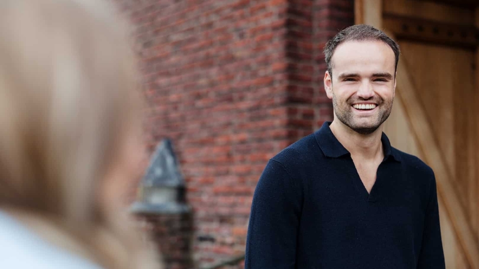 Een man staat buiten voor een bakstenen muur en glimlacht terwijl hij met iemand in gesprek is.