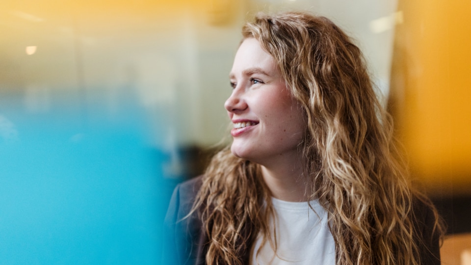 Een vrouw met krullend haar kijkt glimlachend opzij in een kantooromgeving, alsof ze naar een collega luistert.