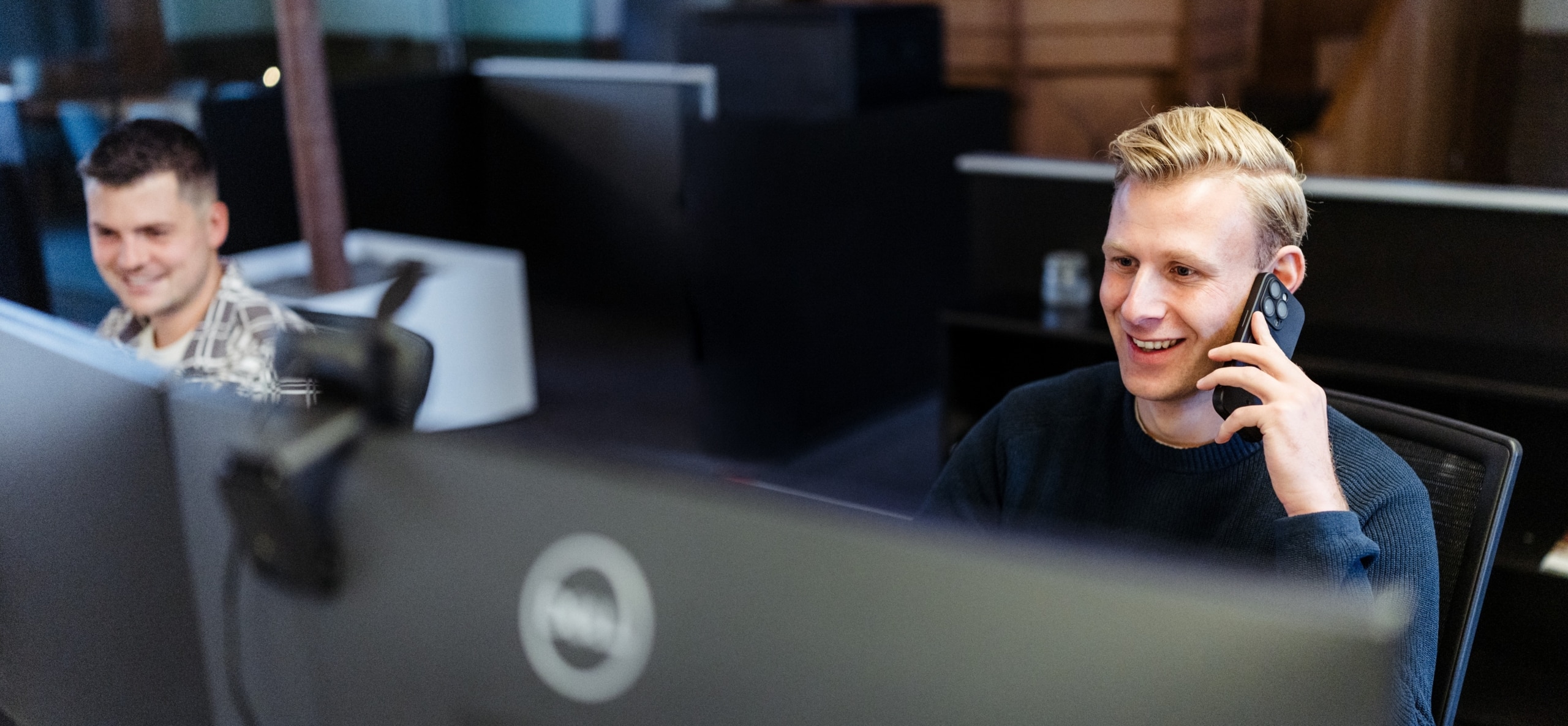 Een man zit achter zijn bureau in een kantoor en belt met zijn smartphone, terwijl een collega op de achtergrond achter een computer werkt.