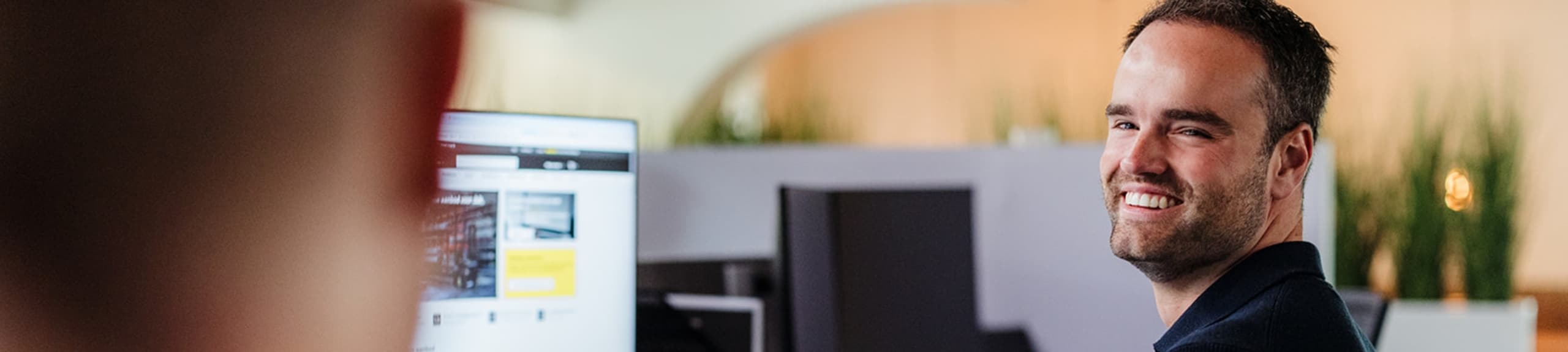 Een man zit achter zijn bureau in een kantoor en kijkt glimlachend op van zijn computerscherm terwijl hij met een collega praat.