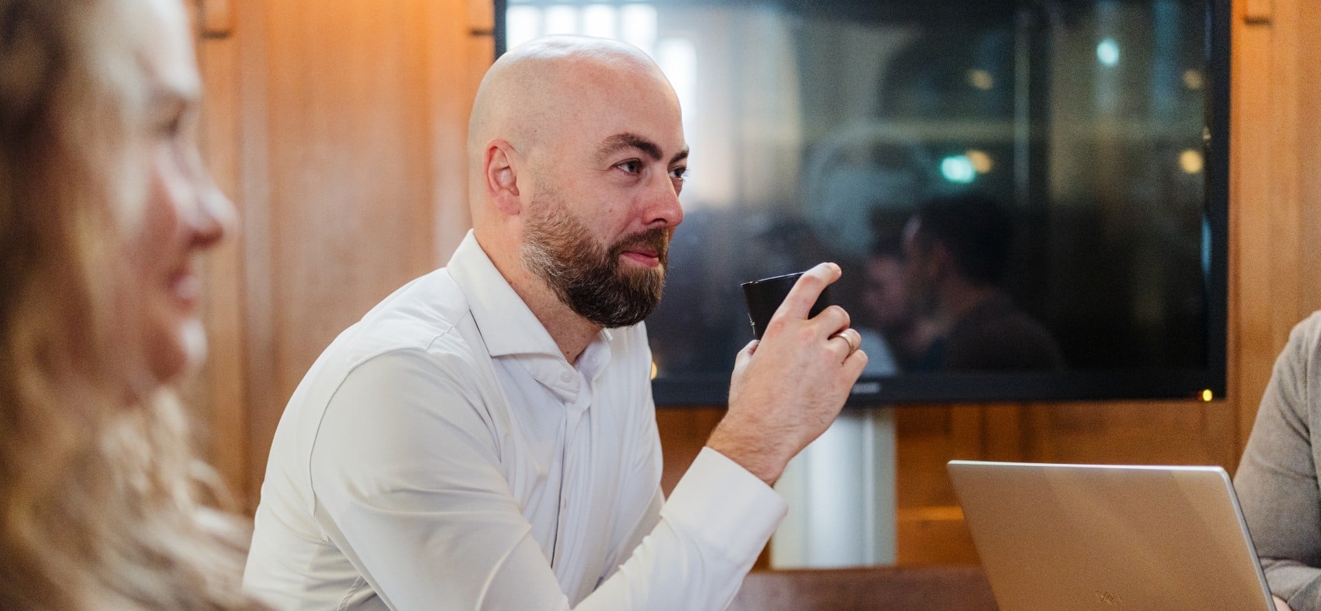 Een medewerker drinkt uit een kopje tijdens een overleg aan tafel met een laptop in een kantooromgeving.