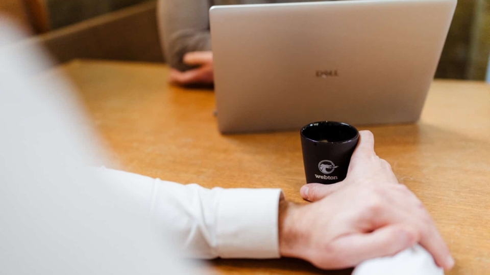 Een close-up van een hand die een zwart kopje vasthoudt, terwijl een laptop op een vergadertafel staat. De persoon in de afbeelding is bezig met een gesprek tijdens een vergadering.