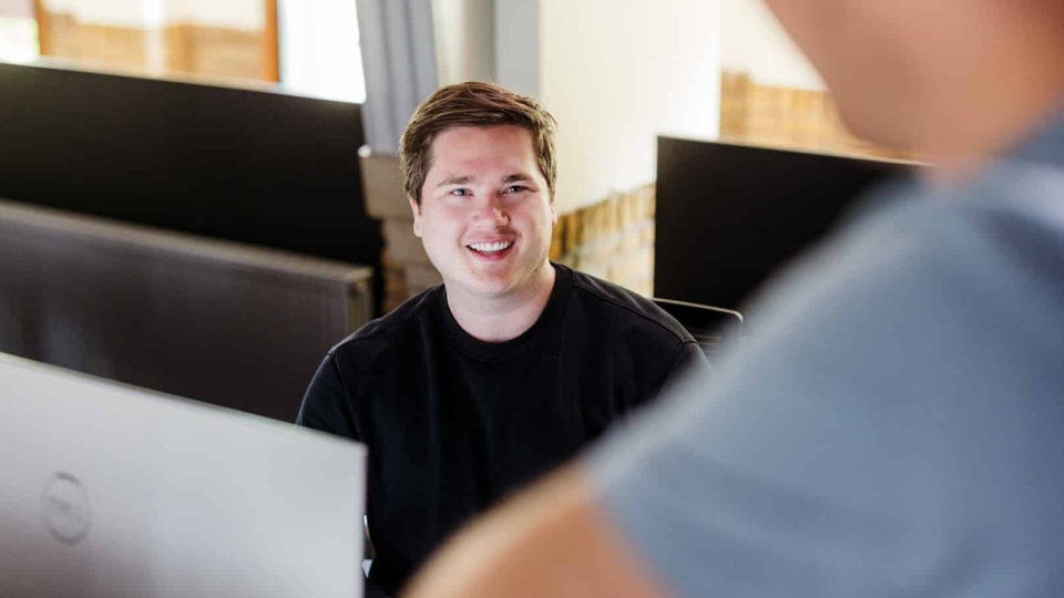 Een man zit achter zijn computer in een kantoor en glimlacht terwijl hij in gesprek is met een collega.
