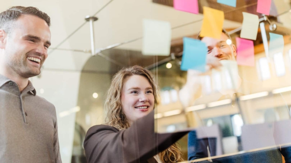 Drie collega’s plakken gekleurde post-its op een glazen wand tijdens een brainstorm en lachen samen.