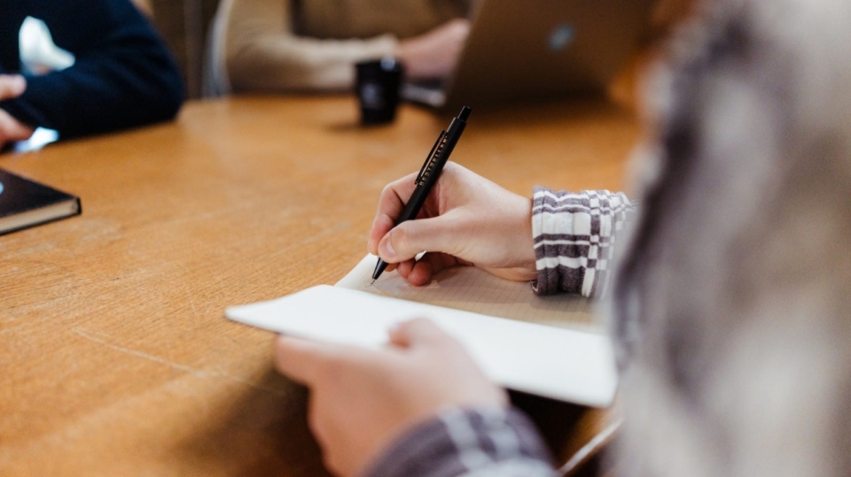Close-up van een hand die aantekeningen maakt in een notitieblok tijdens een vergadering, met een laptop op tafel op de achtergrond.