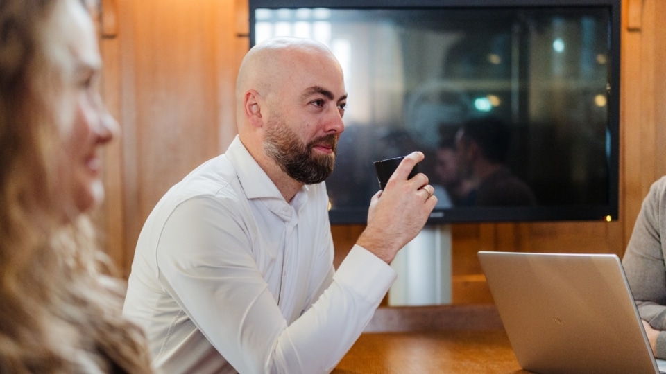 Een man zit aan een vergadertafel met een kop koffie in zijn hand tijdens een overleg, met een laptop op tafel.