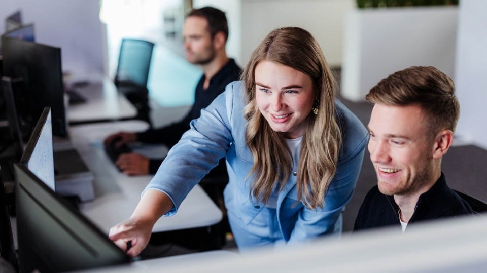 Een vrouw wijst enthousiast naar het scherm van de computer terwijl ze met een collega samenwerkt. Beide lachen terwijl ze naar het scherm kijken. Op de achtergrond is een andere collega bezig met zijn eigen werk.
