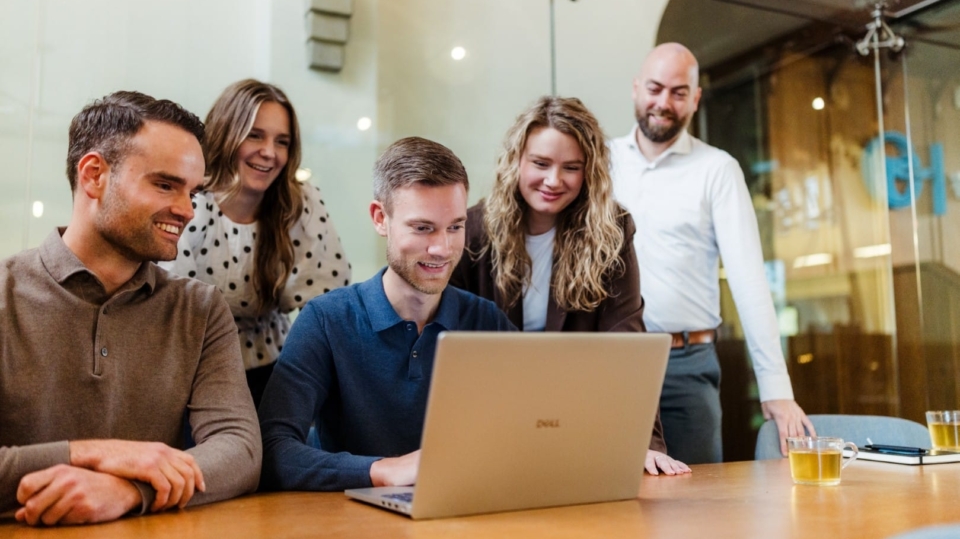 Vijf mensen staan rond een laptop die op een tafel staat. Ze kijken naar het scherm met interesse en lachen, terwijl ze samenwerken aan een project. De sfeer is informeel en samenwerkingsgericht.
