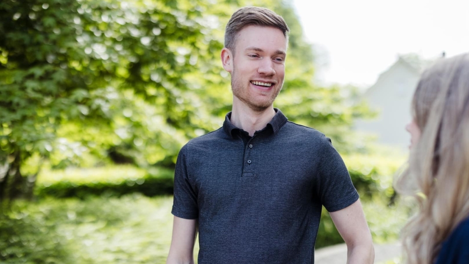 Een man staat buiten en lacht terwijl hij in gesprek is met een collega, met groen en bomen op de achtergrond.