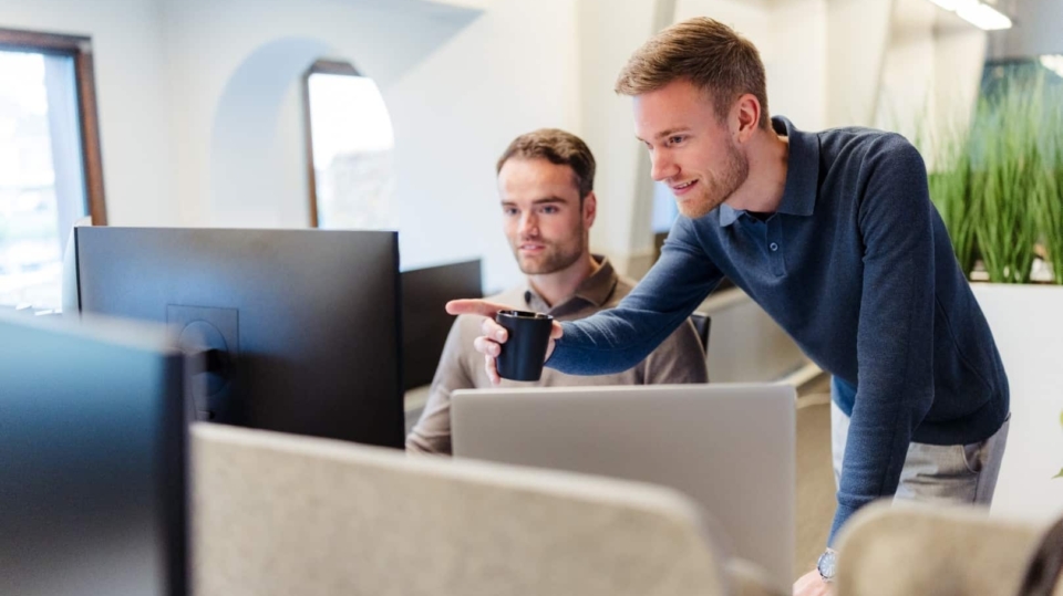 Twee collega’s werken samen achter een bureau en kijken naar een computerscherm, waarbij één man naar het scherm wijst.