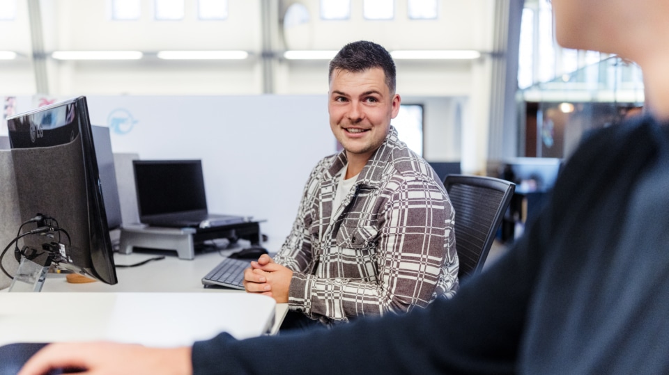 Een man glimlacht terwijl hij naar een collega kijkt, die zich naast zijn bureau bevindt. Hij heeft een lichte glimlach en kijkt geïnteresseerd naar de persoon aan de andere kant van zijn bureau.