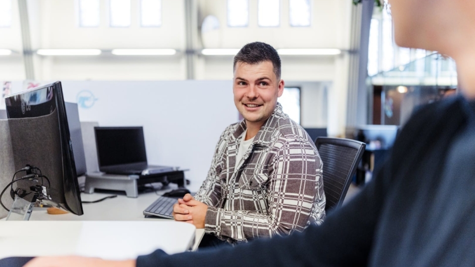 Een man zit achter zijn bureau in een kantoor en kijkt glimlachend naar een collega tijdens het werk.