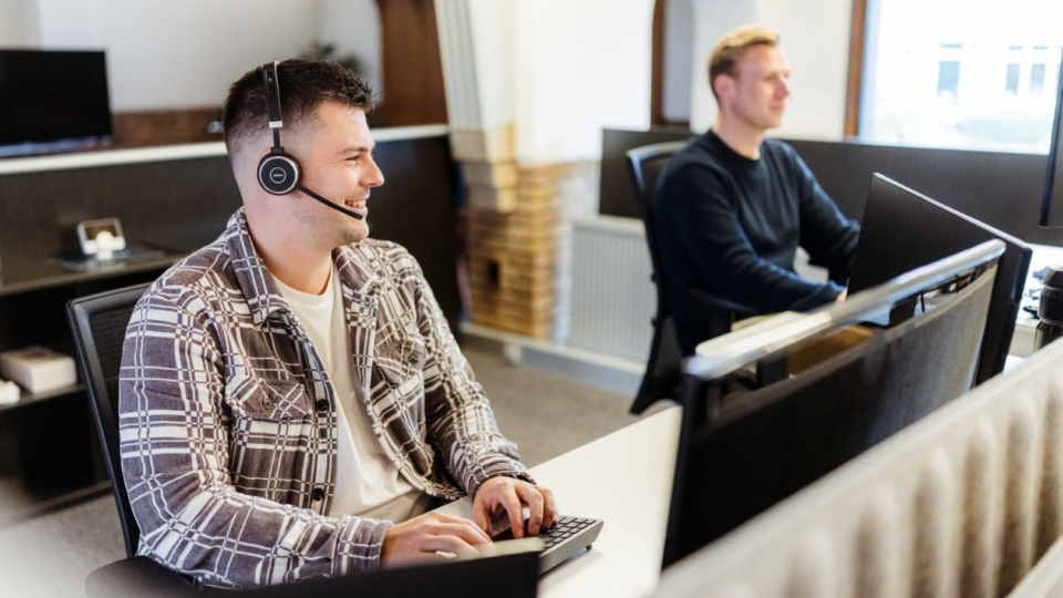 Een man met headset werkt achter zijn computer in een kantooromgeving, terwijl een collega op de achtergrond ook achter een bureau werkt.