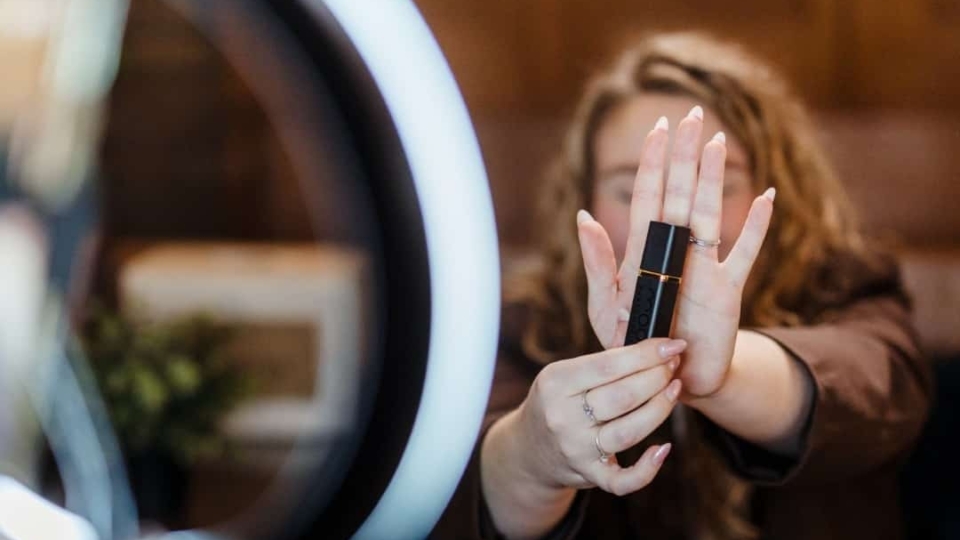 Een vrouw houdt een cosmetisch product voor een ringlamp en smartphonecamera, alsof ze het presenteert voor een video of social media-opname.