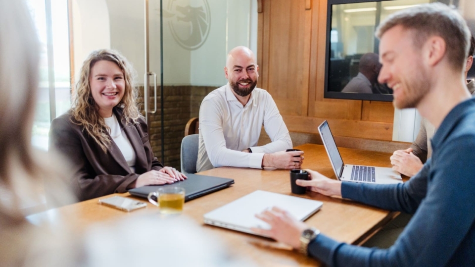 Drie collega’s zitten aan een vergadertafel met laptops en notitieblokken en lachen tijdens een informeel overleg.