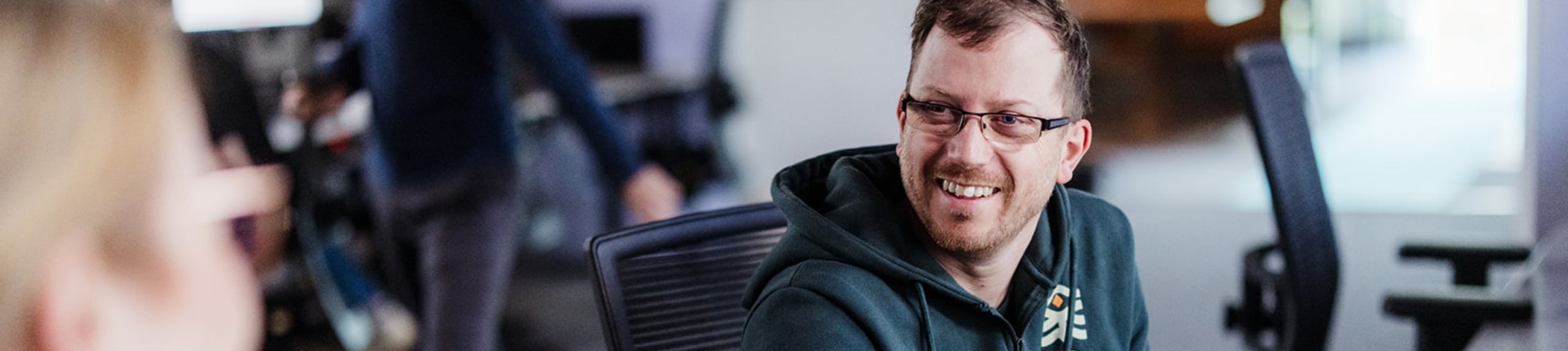 Een man zit achter zijn bureau in een kantoor en glimlacht naar een collega terwijl hij aan zijn computer werkt.